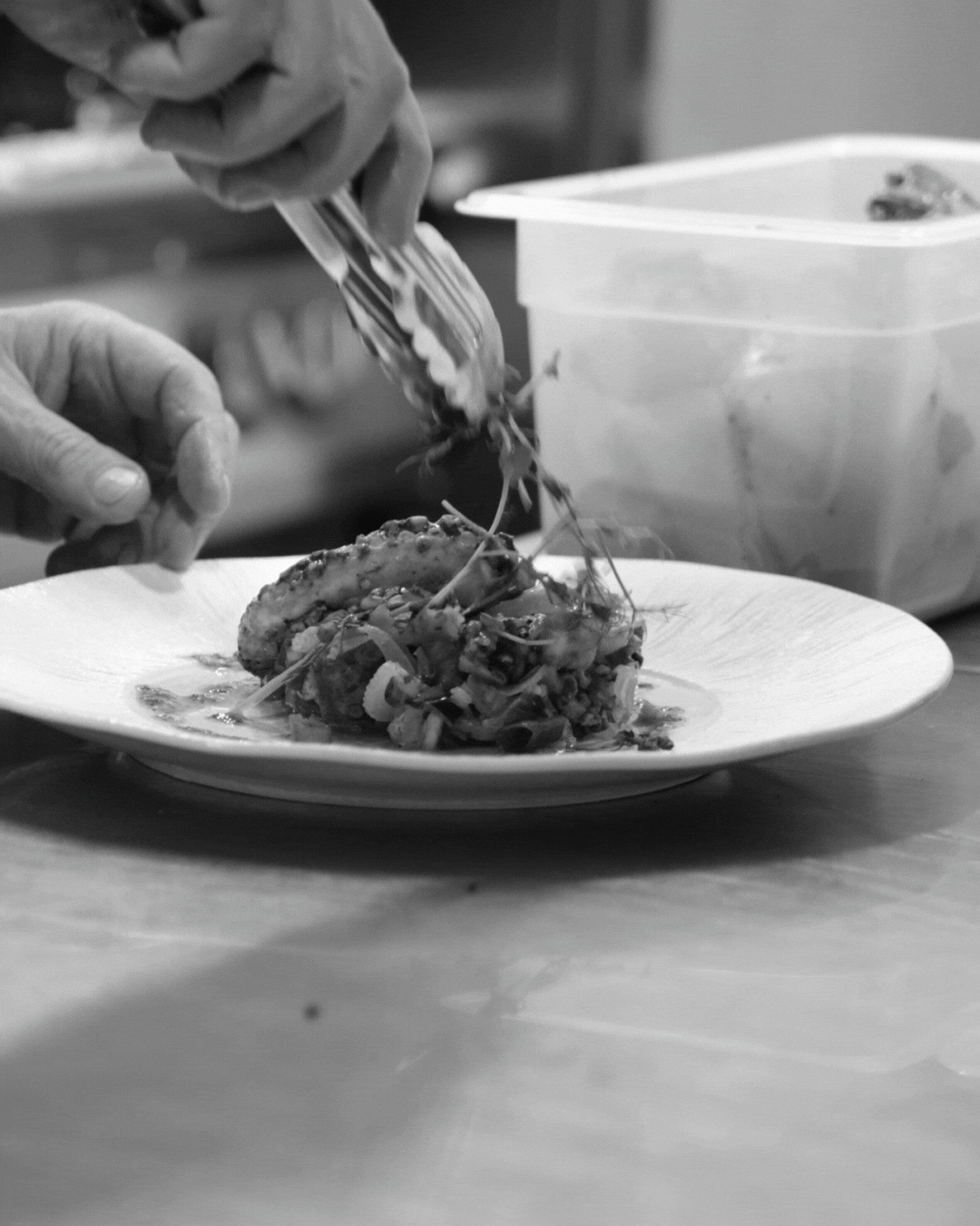 Une personne prépare un plat dans une cuisine, en utilisant des gants en plastique, avec un plat blanc contenant de la nourriture, et une boîte en plastique transparente en arrière-plan.