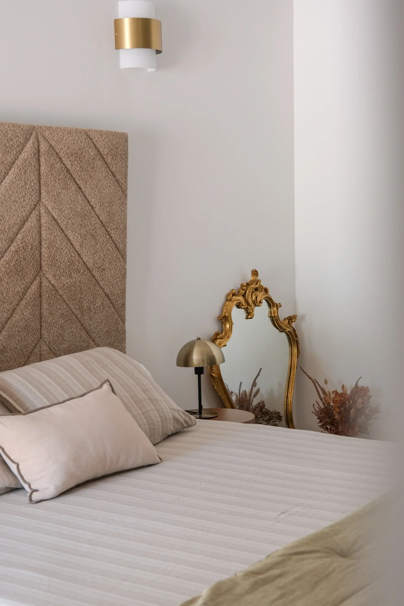 Chambre élégante avec lit à drap blanc, plusieurs coussins beige, tête de lit en tissu beige, lampe de chevet en métal, miroir doré de style baroque, décoration florale sèche dans un vase, mur blanc