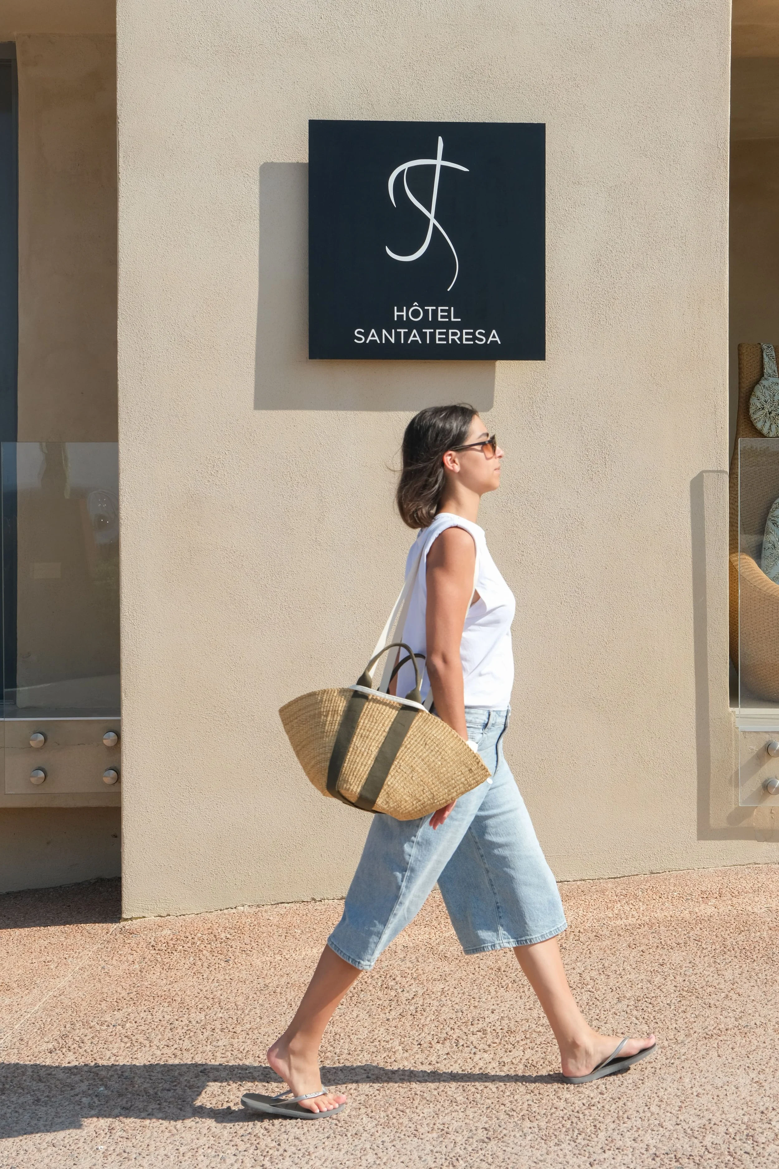 Une personne qui marche devant un hôtel, avec un haut blanc, un bermuda en jean bleu, des tongs, un panier en osier avec des poignées en tissu vert, et des lunettes.