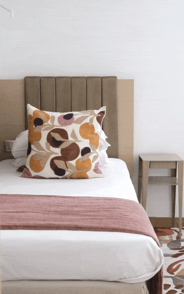 Lit avec un oreiller à motifs abstraits et une couverture rose en tissu à carreaux, dans une chambre minimale avec un mur blanc et une table de nuit en bois.