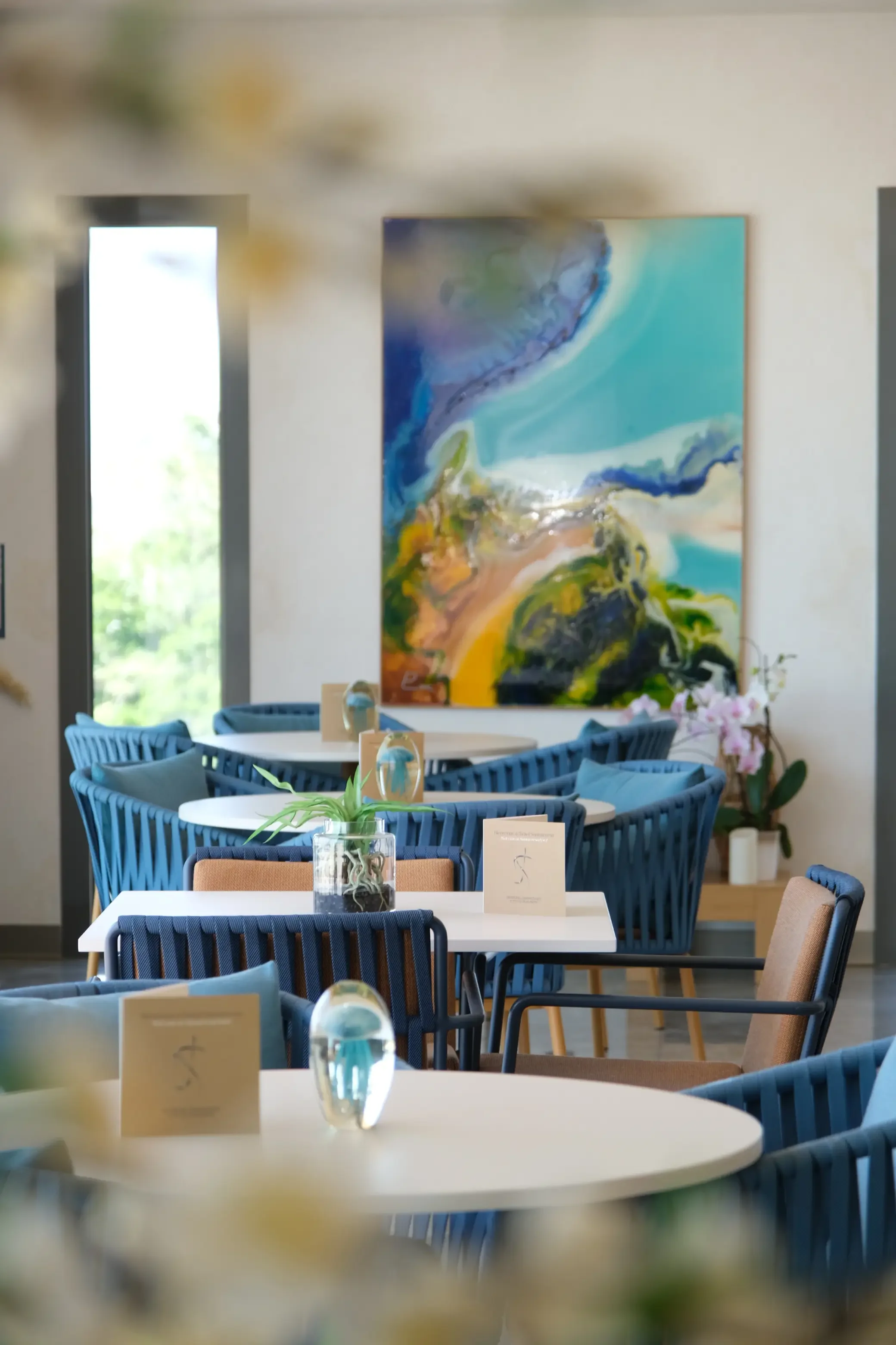 Salle à manger avec des tables rondes, des chaises bleues, un tableau abstrait coloré sur le mur et des plantes en décoration.