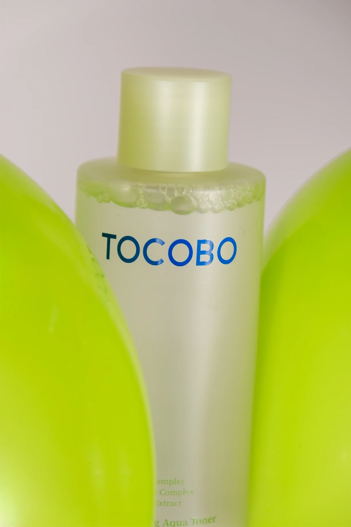Une bouteille blanche avec le nom 'TOCOBO' en lettres bleues, entourée de deux objets verts flous, probablement des produits de soin ou de beauté.