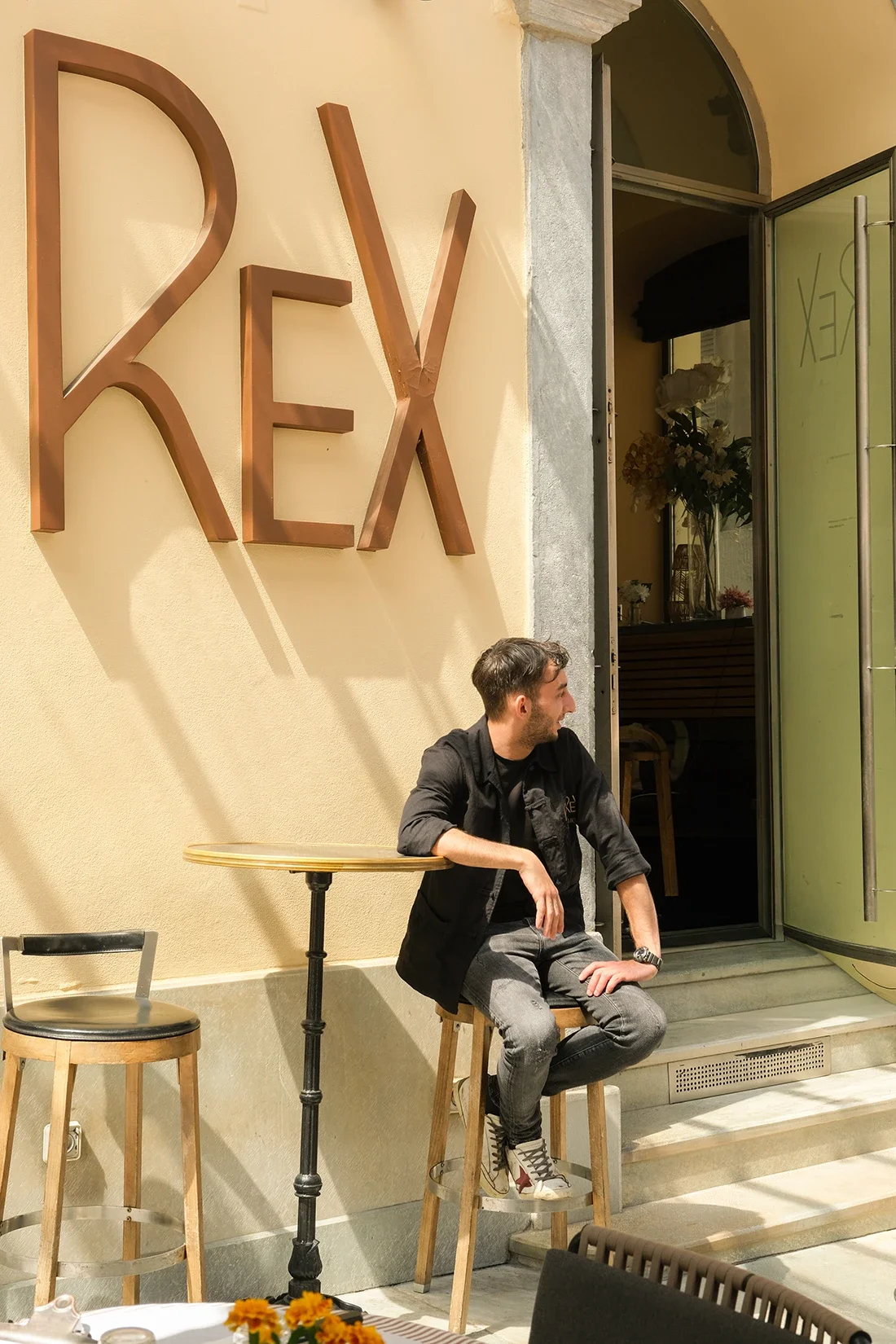 Un homme assis sur un tabouret en bois à l'extérieur d'un café ou restaurant, devant une porte ouverte, avec une grande enseigne en métal blanche et lettres en cuivre qui dit 'REX' sur un mur jaune clair.