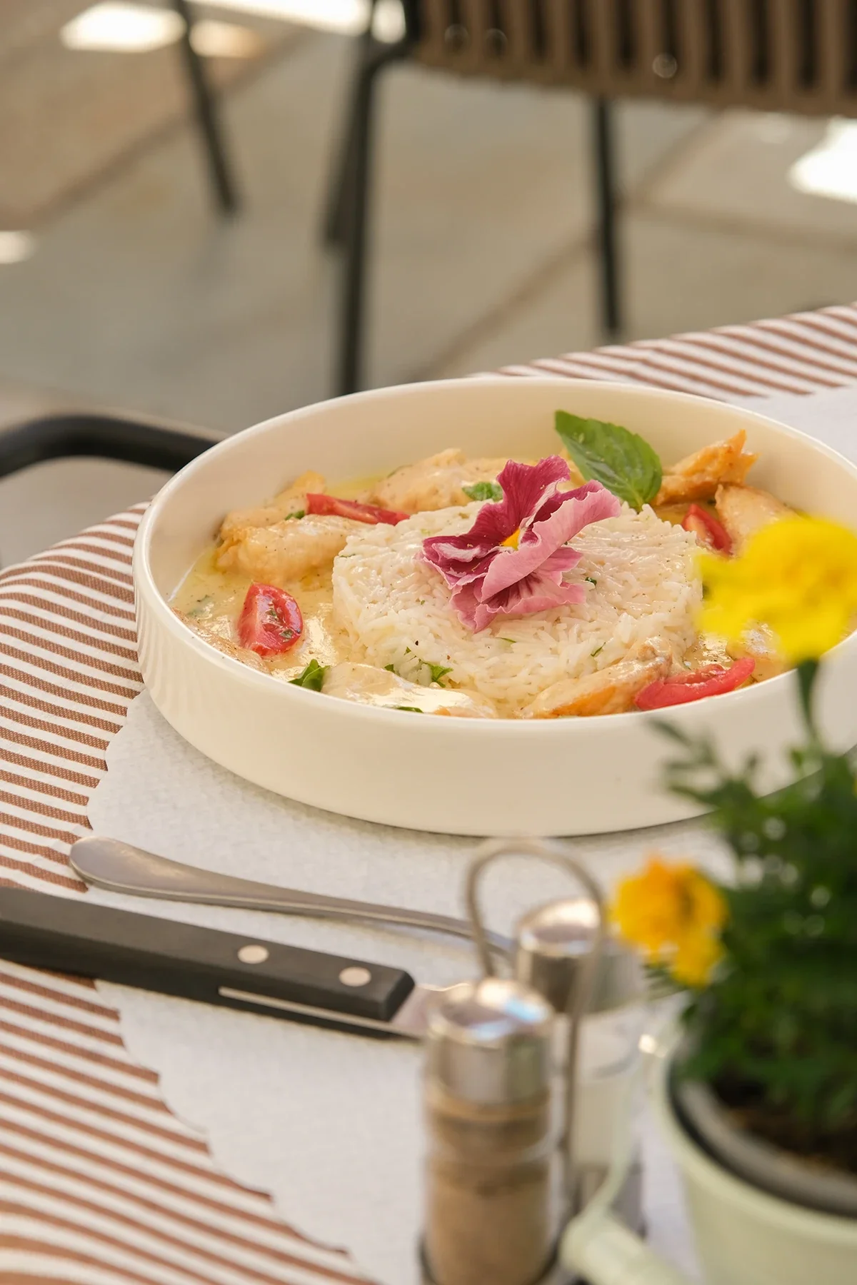 Un plat de riz avec du poulet, des tomates, une feuille de menthe, une fleur rose décorative, dans un bol blanc, placé sur une table avec une nappe à rayures marron et blanche, avec des ustensiles et un vase avec des fleurs en premier plan.