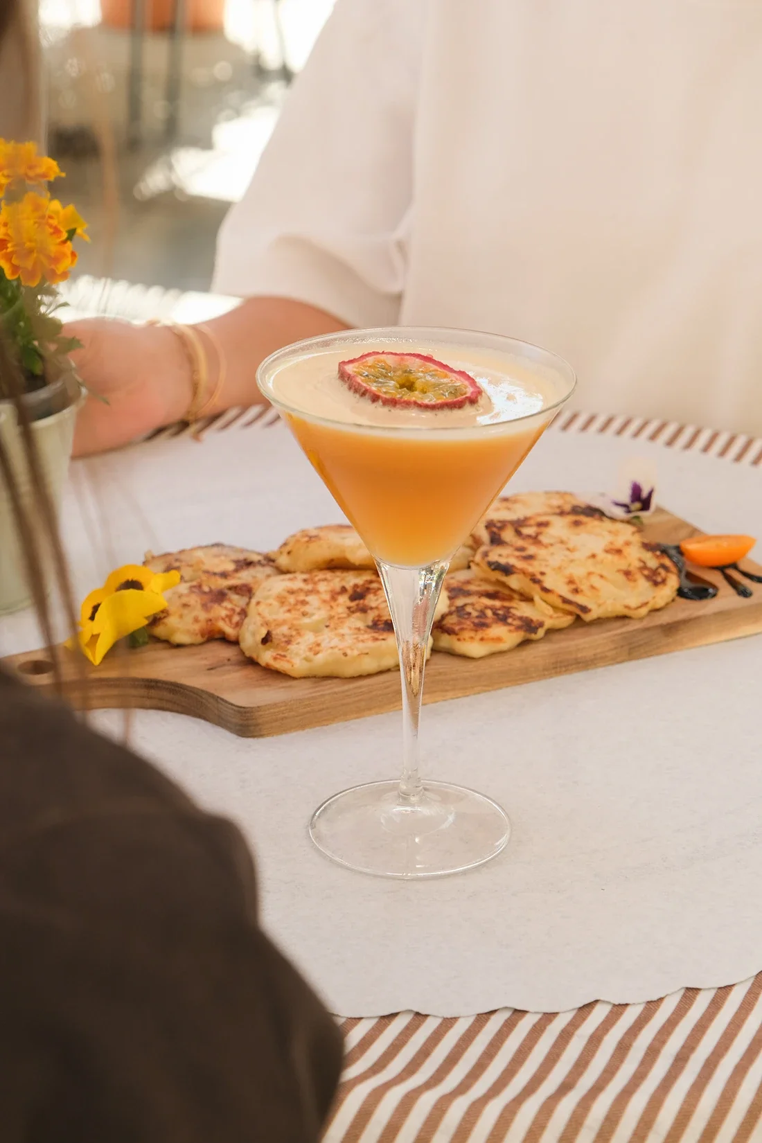 Cocktail orange avec tranche de fruit sur le dessus, accompagné de galettes de pommes de terre ou pancakes, sur une planche en bois, avec fleurs décoratives, dans un cadre bien éclairé et convivial.