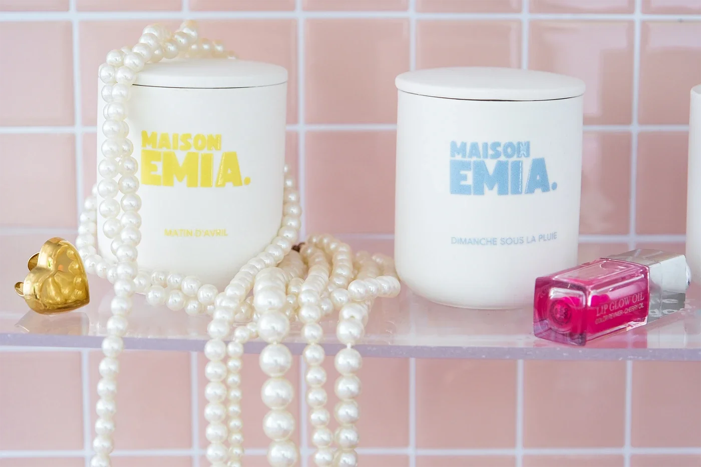 Deux bocaux en céramique blancs avec des inscriptions jaunes et bleues, une chaîne de perles blanches, un anneau en or en forme de cœur, et un vernis à ongles rose sur une étagère en verre sur un mur de carreaux roses.
