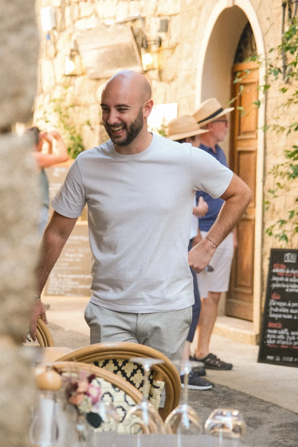 Un homme souriant portant un t-shirt blanc et un pantalon clair, se tenant dans une ambiance de restaurant en extérieur, avec des personnes portant des chapeaux en arrière-plan.