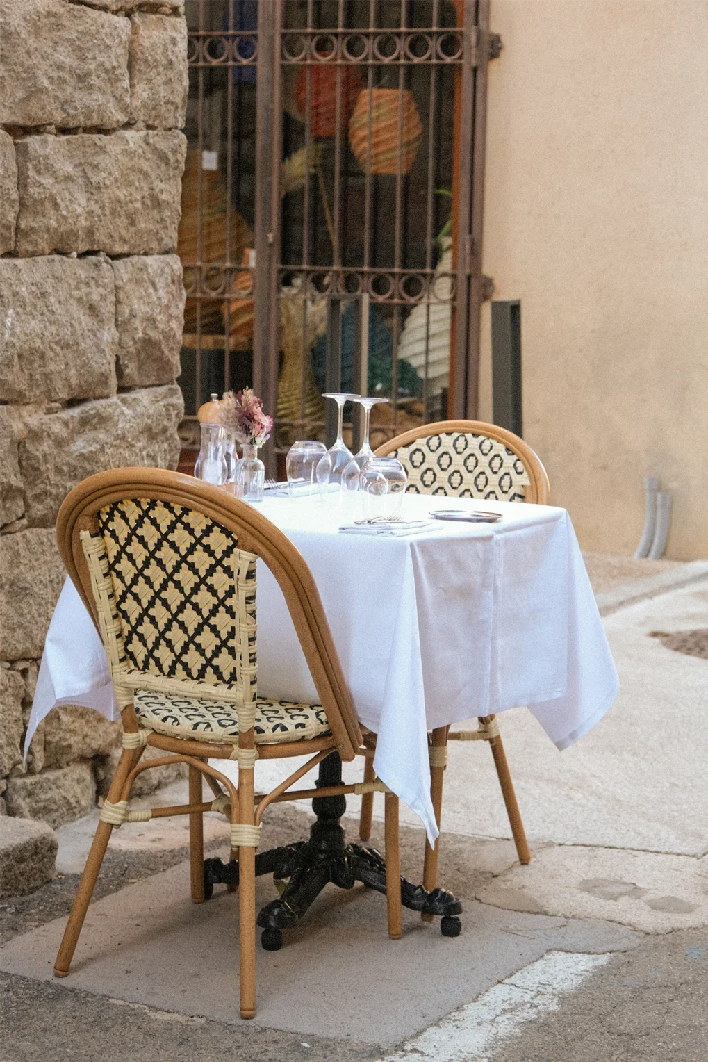 Table dressée pour un dîner, avec une nappe blanche, des couverts, des verres, et une petite décoration florale, situé en extérieur contre un mur en pierre.
