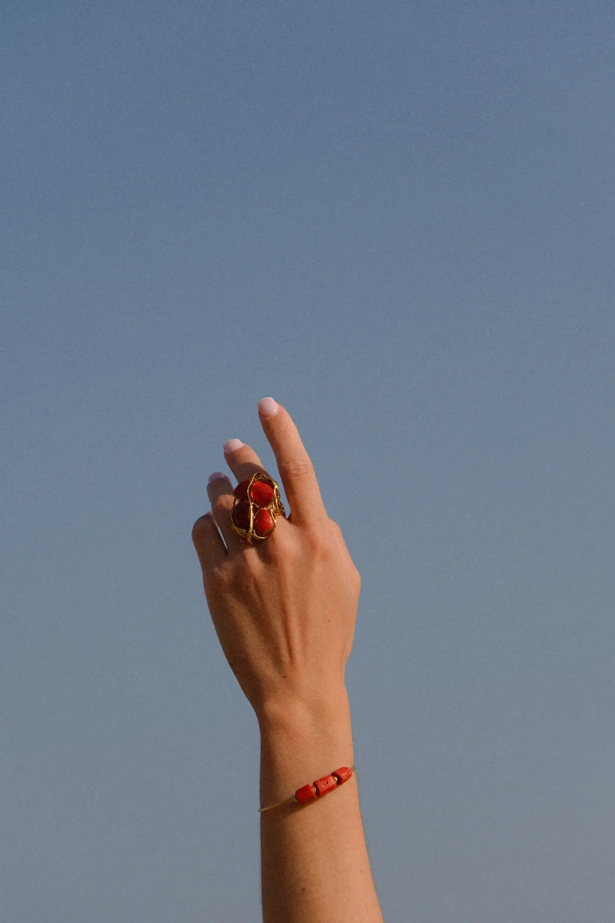 Une main féminine portant plusieurs bijoux rouges, y compris un grand anneau, un bracelet et un collier, levée devant un ciel bleu clair.