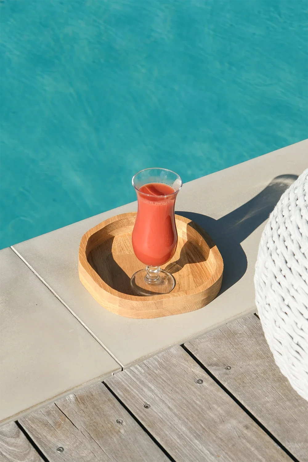 Verre de smoothie rose à côté d'une piscine, placé sur un plateau en bois sur une banquette en bois.