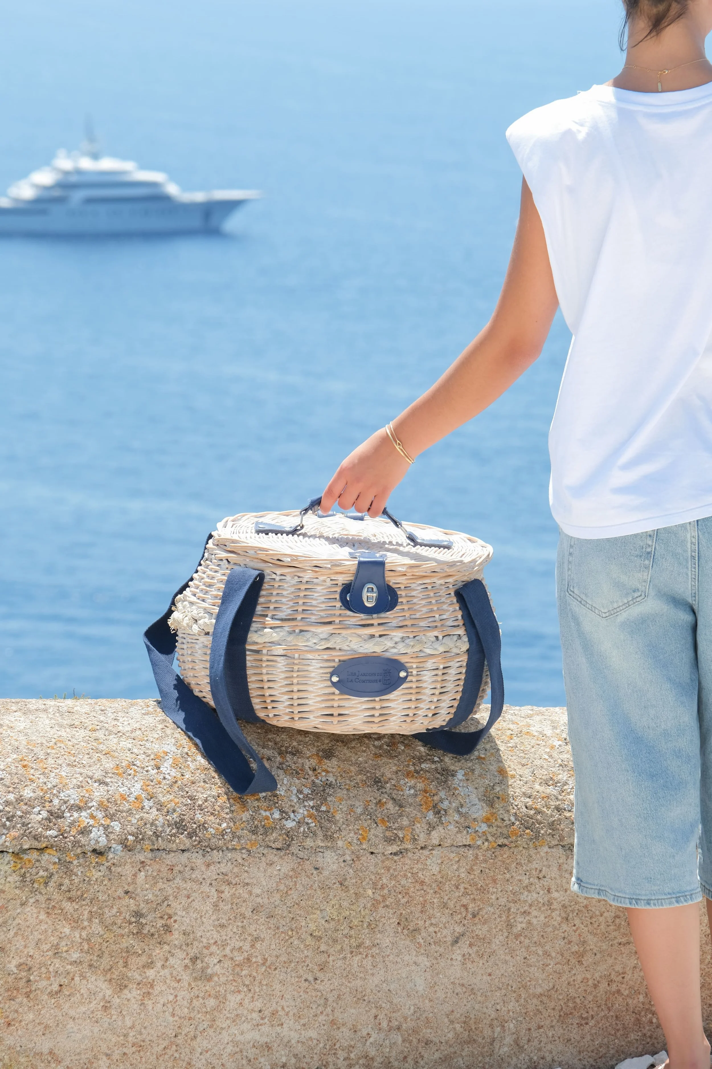 Une main tenant un panier en osier avec des poignées en tissus bleues, posé sur un muret. La personne porte un haut blanc et un bermuda en jean bleu. En fond il y a la mer, et un bateau.
