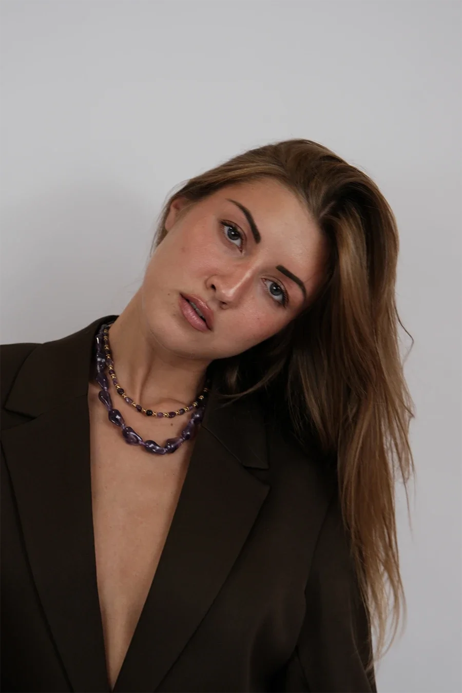 jeune femme aux yeux bleus, cheveux bruns, portant un blazer noir et deux colliers en perles violettes, posé en biais près d'un fond gris clair