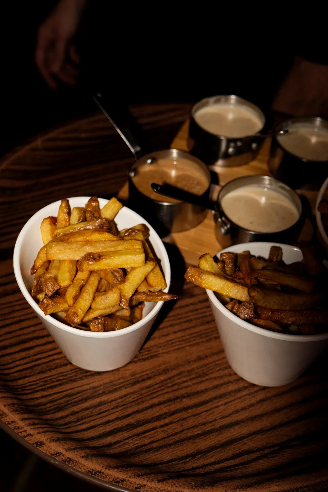 Frites dans deux coupes en papier blanc avec différentes sauces en petits pots en métal à côté.