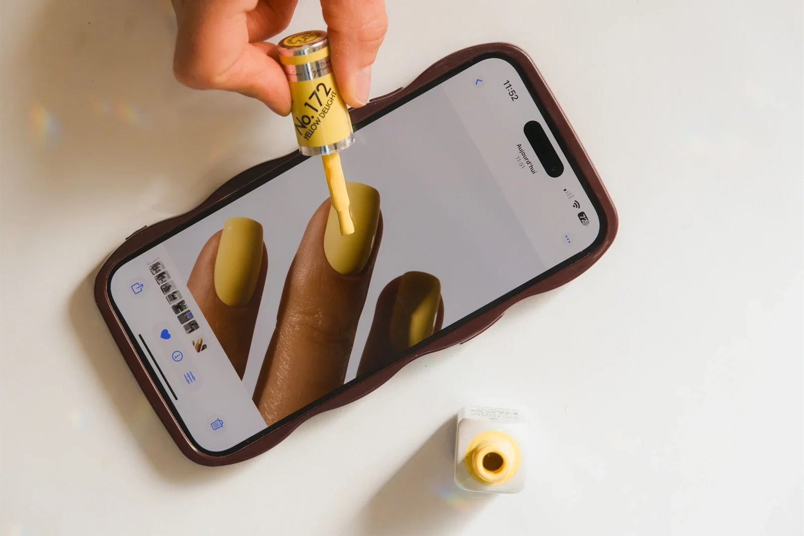 Une main tenant un pinceau de peinture jaune qui est au-dessus d'un téléphone portable affichant une image d'une main avec des ongles jaunes. Le pinceau de peinture est à côté d'une bouteille de peinture jaune.