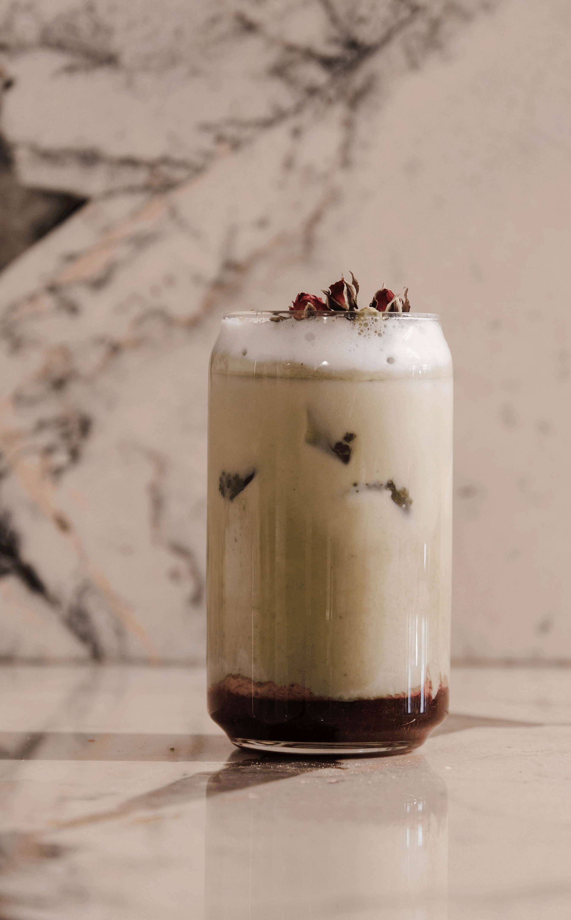 Verre transparent contenant une boisson à plusieurs couches de couleurs : rouge foncé en bas, blanche au milieu, et mousse au sommet, garnie de roses séchées