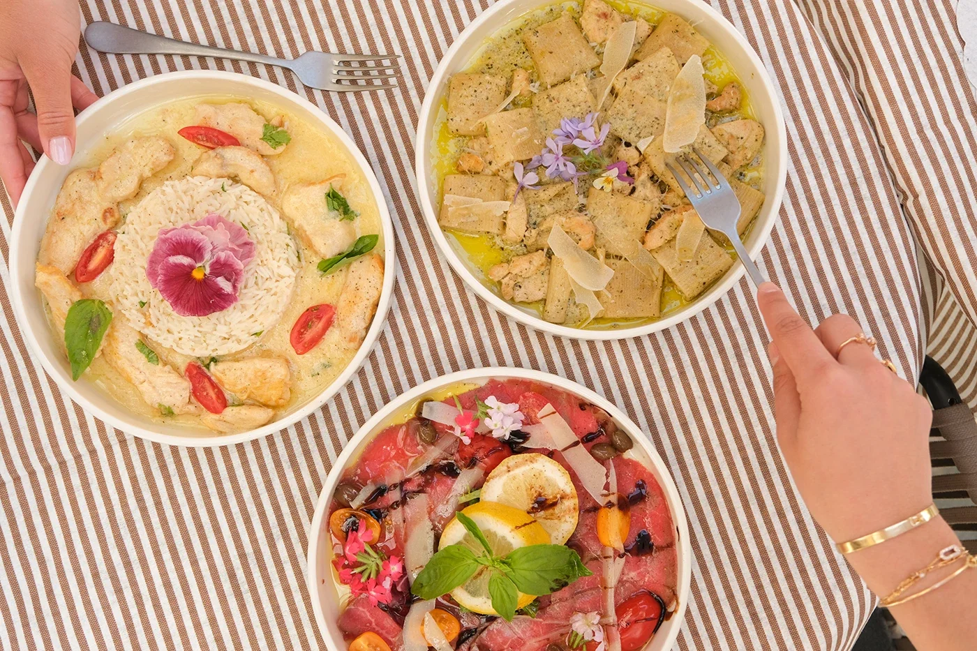 Trois plats de cuisine méditerranéenne ou orientale, présentés sur une nappe à rayures, avec des fleurs comestibles et des légumes frais, dont du riz, du poisson, des légumes et des herbes.