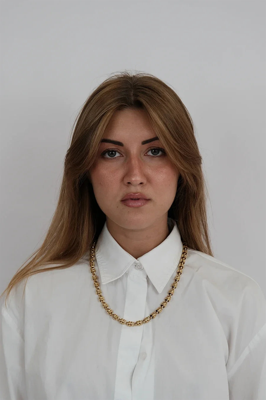 Une femme avec les cheveux bruns, portant une chemise blanche et un collier doré, pose contre un fond gris clair.