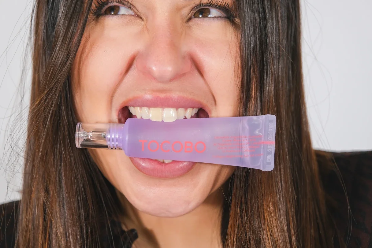 Une femme avec des cheveux bruns tient un tube de gel dans sa bouche, portant une expression de surprise ou de jeu.