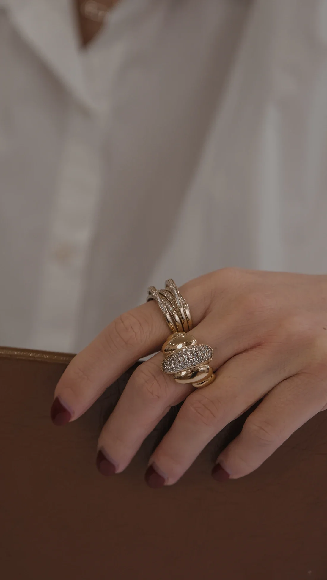 Une main portant plusieurs bagues en or, dont certaines ornées de diamants, posée sur une surface en bois. En arrière-plan, une personne porte une chemise blanche.