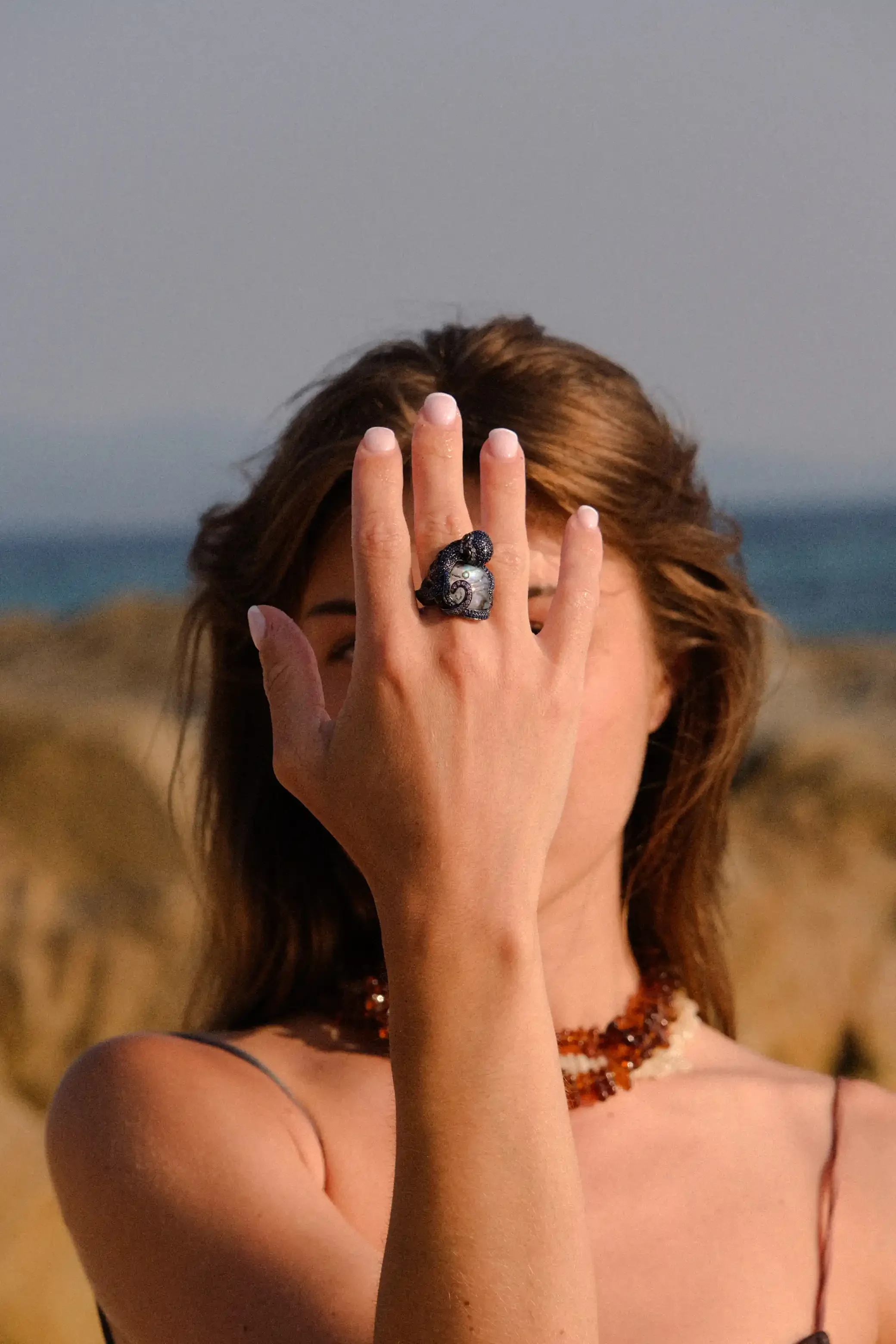 Une femme portant un collier en perles ornée d'une grosse bague noire en forme de serpent, couvrant une partie de son visage avec sa main, devant un paysage en plein air.