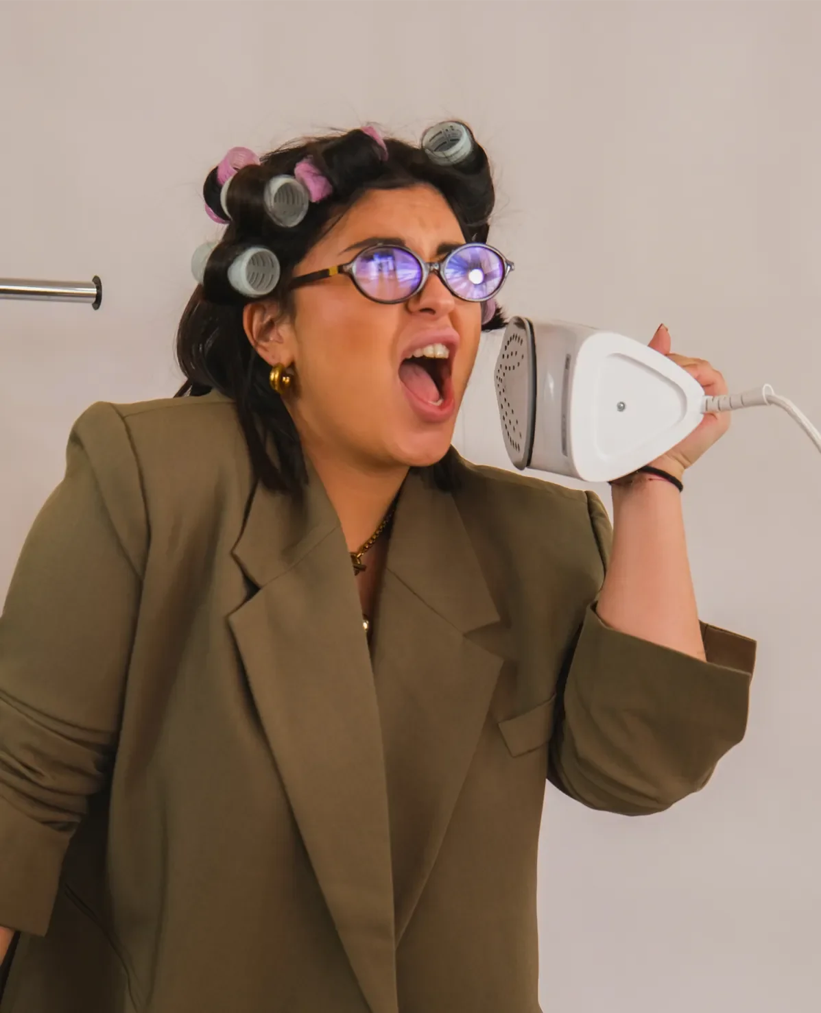 Femme avec rouleaux capillaires dans les cheveux, portant des lunettes et un blazer, tenant un fer à repasser comme un téléphone, semblant faire semblant d'être surprise ou choquée.