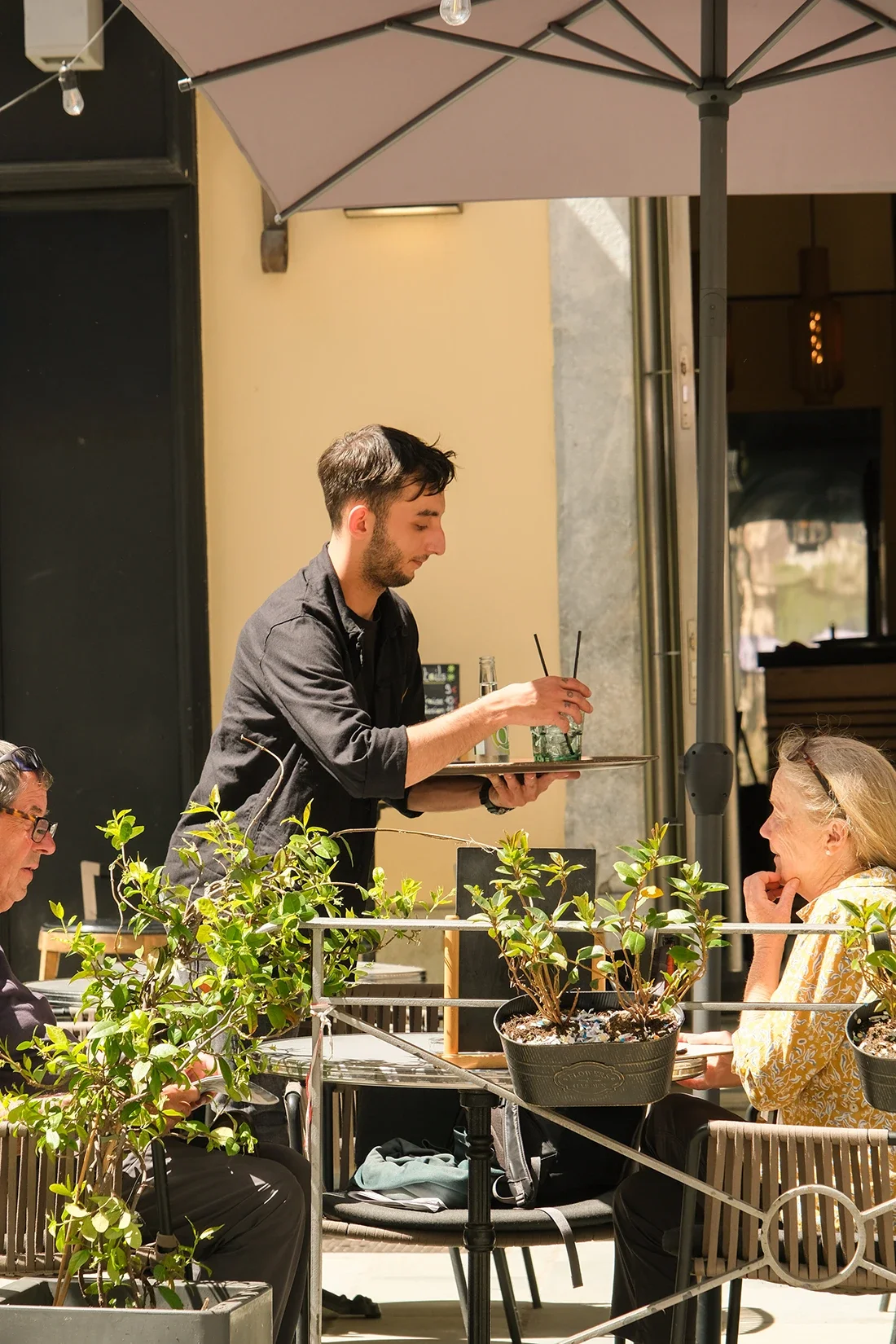 Un serveur sert des boissons à deux clients assis à une table en terrasse d'un café en plein air, avec un parasol et des plantes décoratives autour.