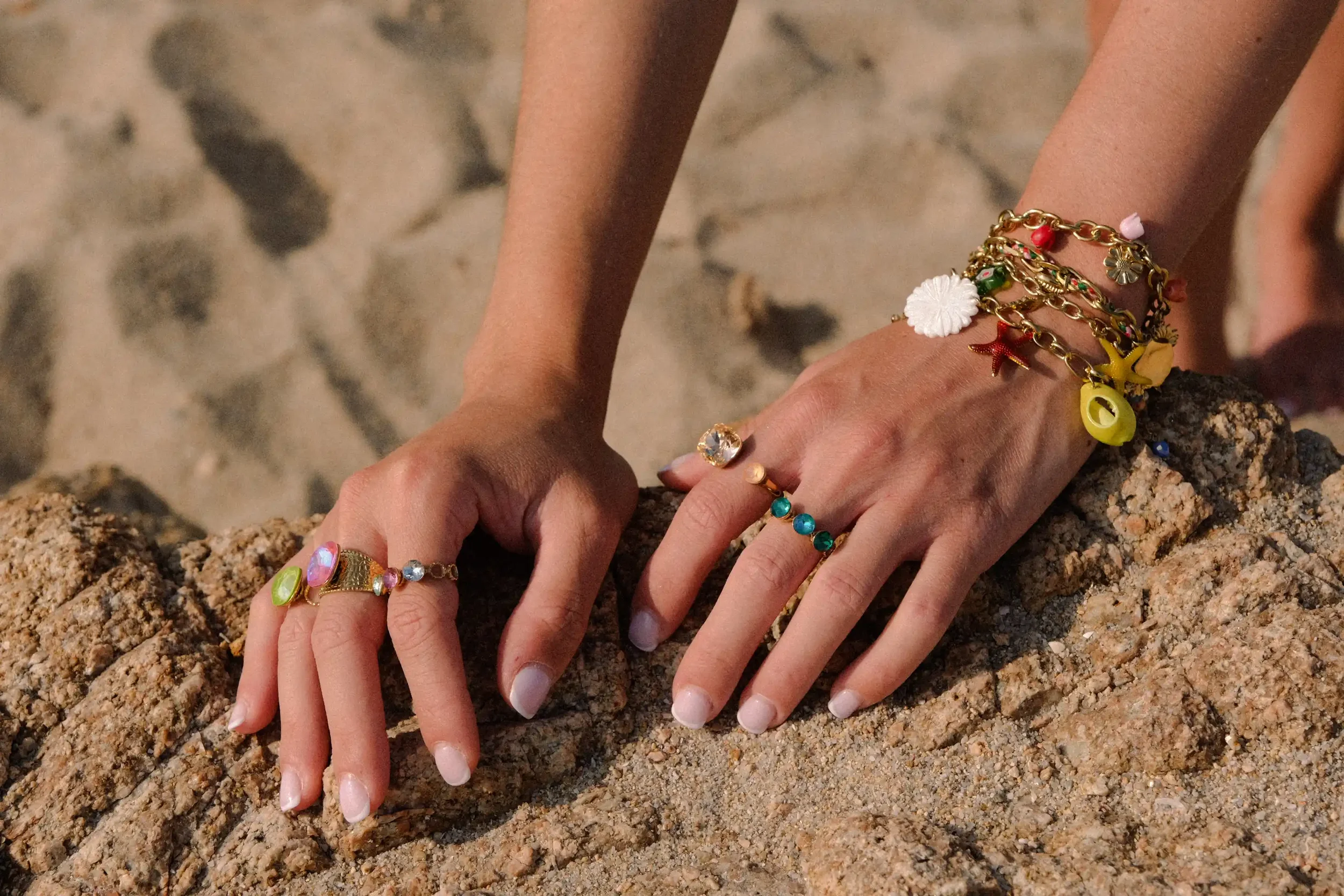 Main nue aux doigts ornés de plusieurs bracelets colorés, posant sur du sable avec un fond légèrement flou