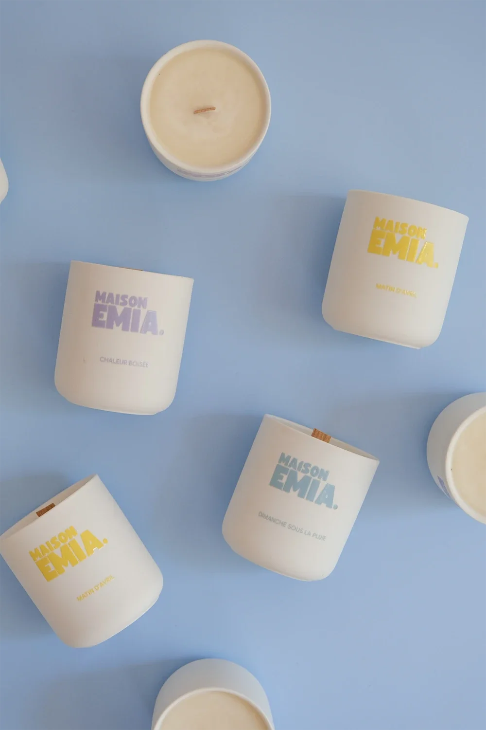 Bougies parfumées dans des tasses blanches avec différentes inscriptions colorées, disposées sur un fond bleu.