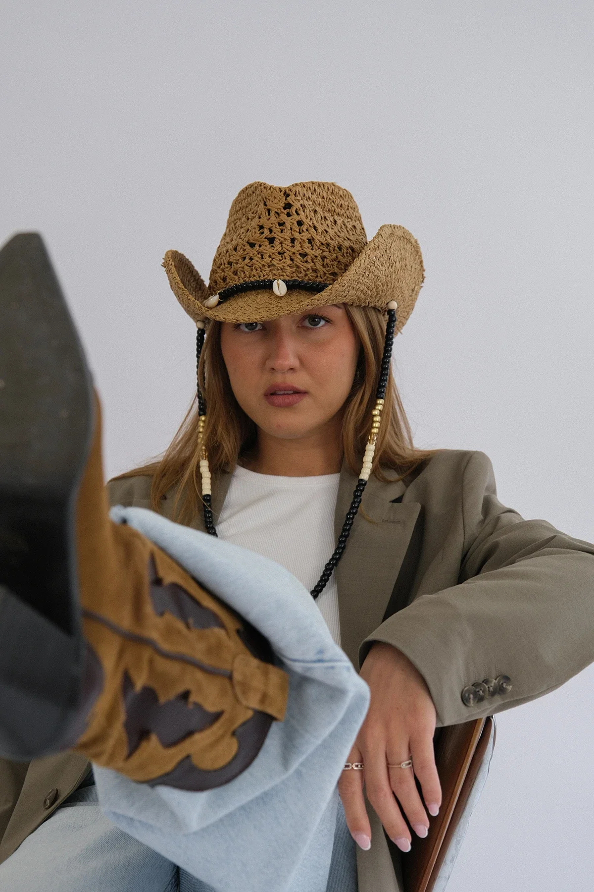 Femme portant un chapeau de cowboy en paille, veste beige, et tenant une chaussure de cowboy