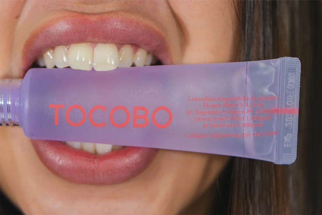 Une personne tenant un tube de crème pour les yeux devant ses dents, avec les lèvres légèrement ouvertes.