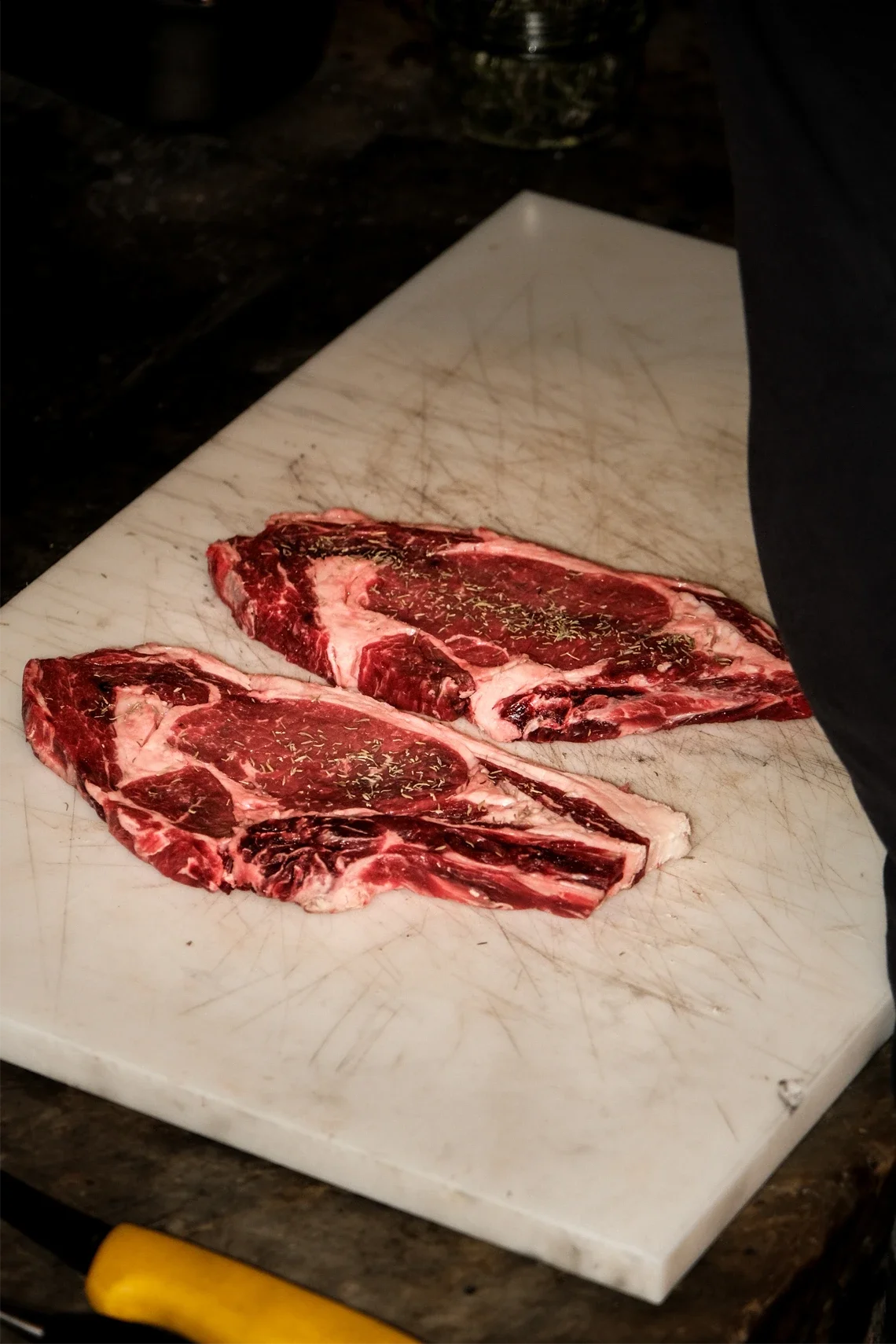 Deux steaks de viande crue assaisonnés avec des herbes, posés sur une planche à découper blanche