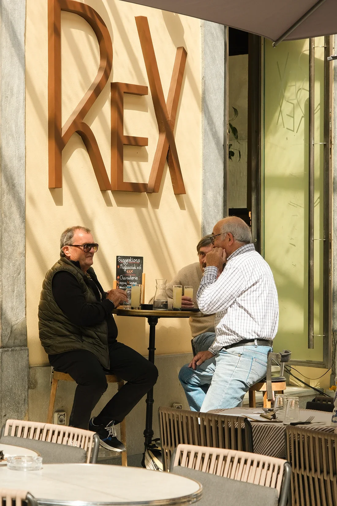Trois hommes assis autour d'une table de café en extérieur, discutant et ayant des boissons. Un panneau propose des suggestions de plats.