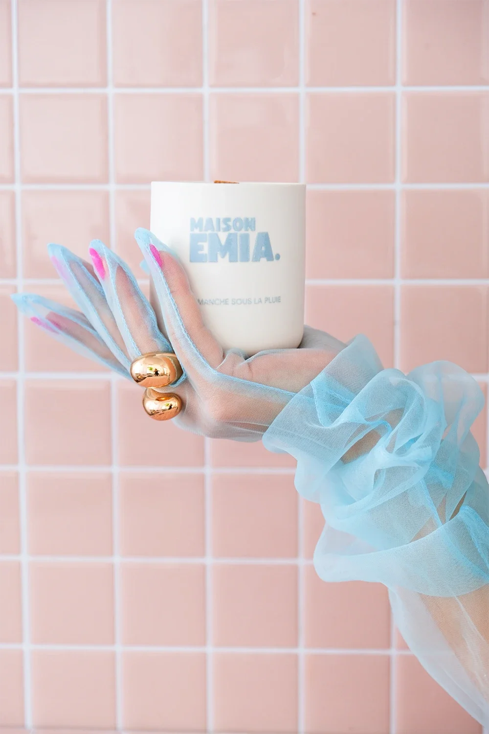 Une main portant un gant en tulle bleu clair tient une tasse blanche avec le texte «MAISON EMIE» en bleu ciel, avec un fond de carreaux roses. La main porte deux gros anneaux dorés.