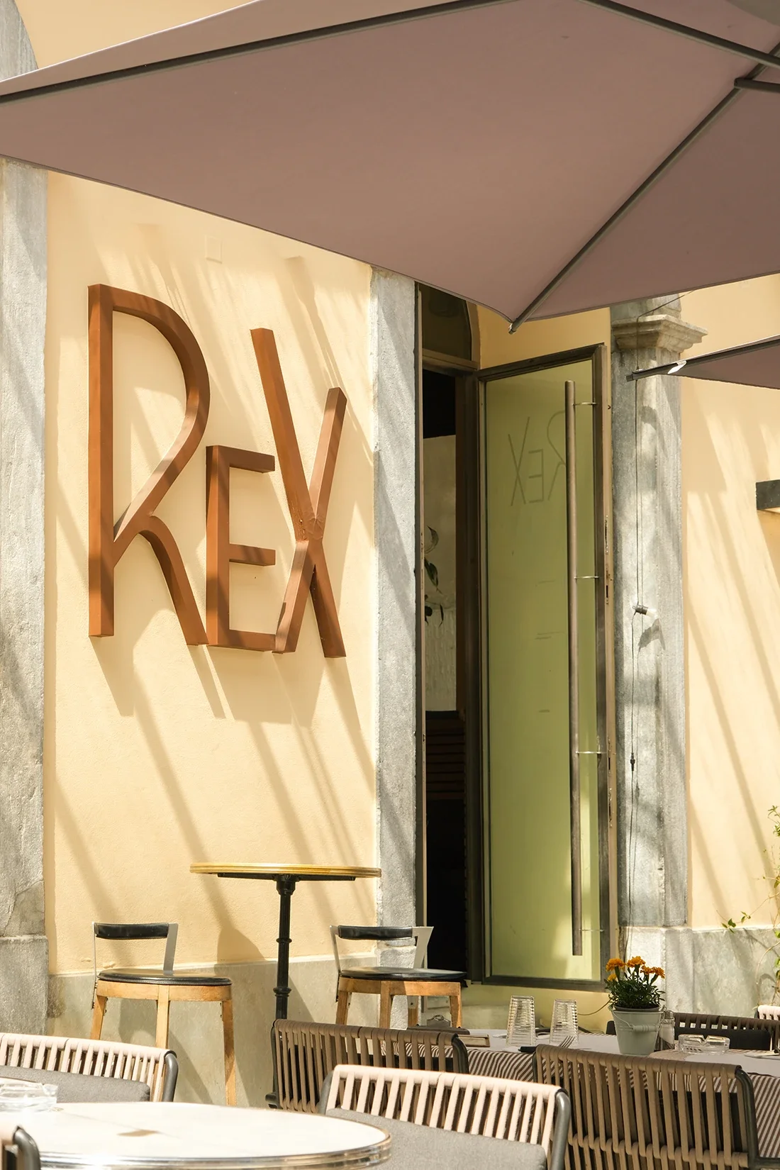 Façade d'un restaurant avec un grand panneau en métal indiquant 'REX', entouré de murs beige, avec une grande porte semi-vitrée ouverte, des tables et chaises en extérieur.