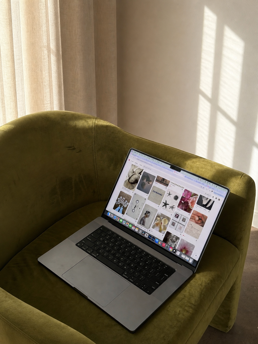 Ordinateur portable ouvert sur une page d'images ou de Pinterest, posé sur un canapé vert en velours dans une pièce lumineuse avec des rideaux blancs laissant passer la lumière
