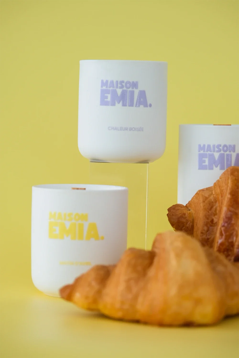 Trois bougies parfumées de la marque Maison Emilia avec un fond jaune et quelques croissants en premier plan.