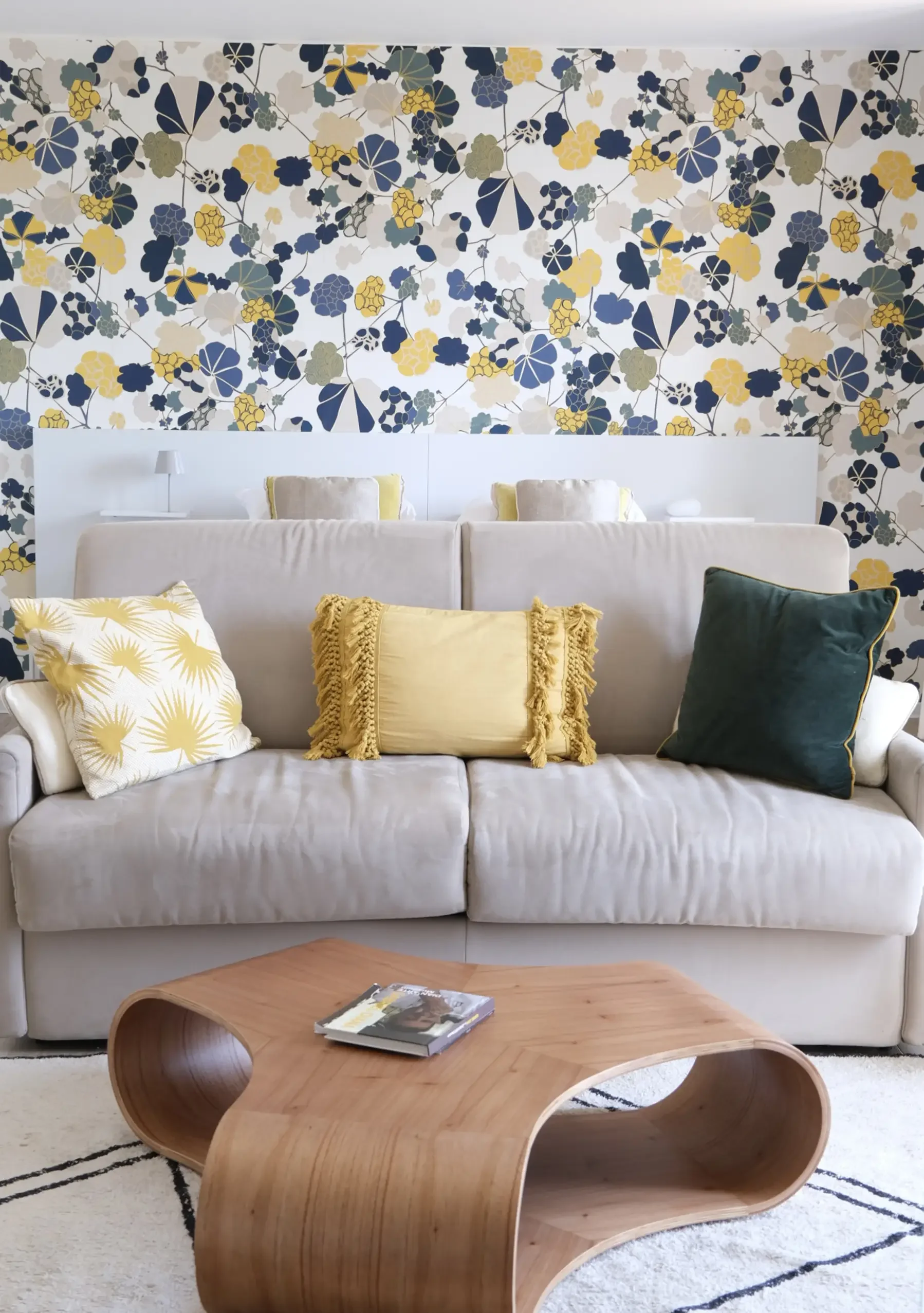 Salon avec un canapé beige, des coussins jaunes et verts, une table en bois moderne, un mur avec un papier peint floral bleu, beige, gris et jaune, et une étagère blanche derrière le canapé.