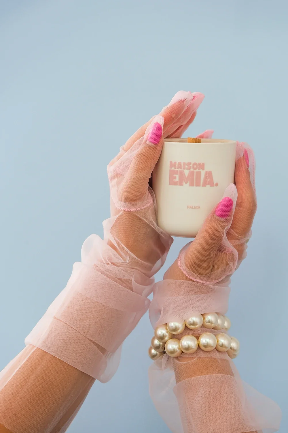 Des mains portant des gants en tulle transparent avec des ongles en rose pâle, tenant une bougie blanche avec l'inscription 'MAISON EMIA' et 'PALMA', contre un fond bleu clair.