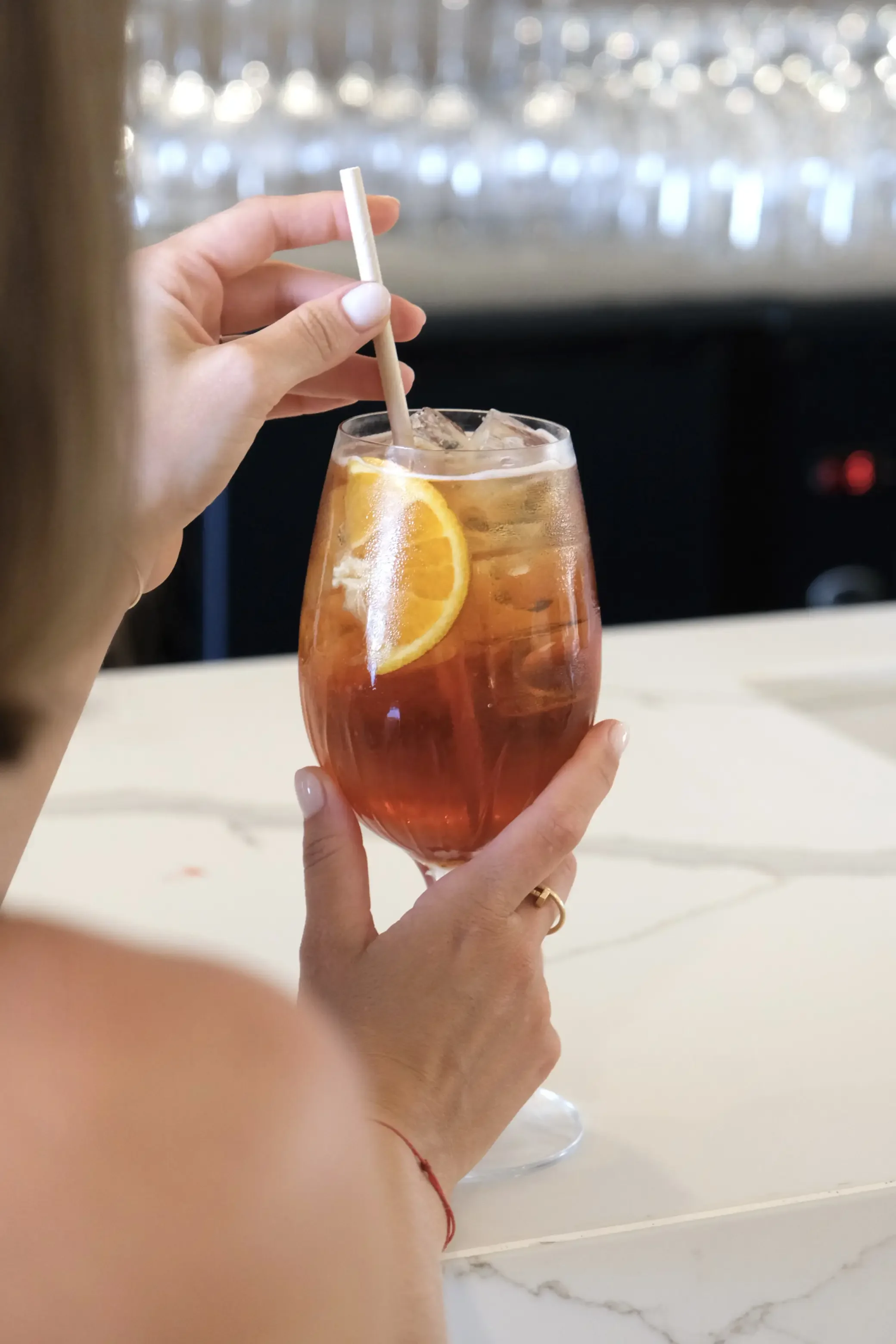 Une personne tient un verre de boisson contenant des glaçons et une tranche de citron, avec une paille en plastique, dans un environnement intérieur.