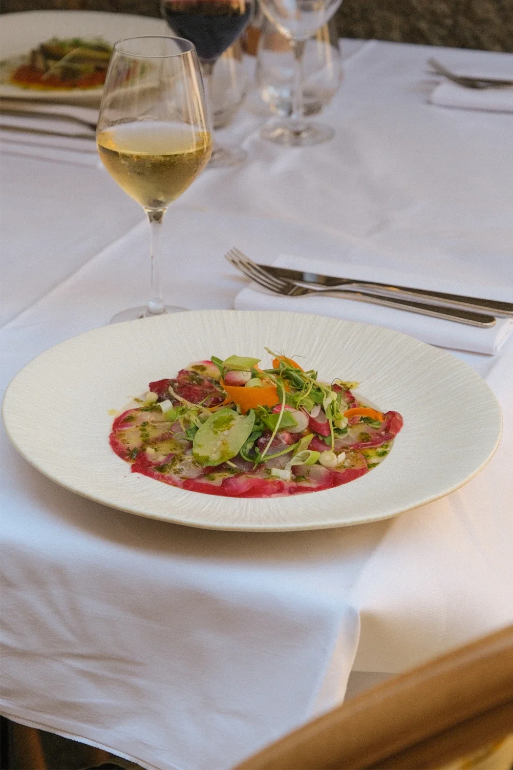 Assiette de carpaccio de veau garnie de microgreens, radis, et légumes finement tranchés, accompagnée d'un verre de vin blanc.