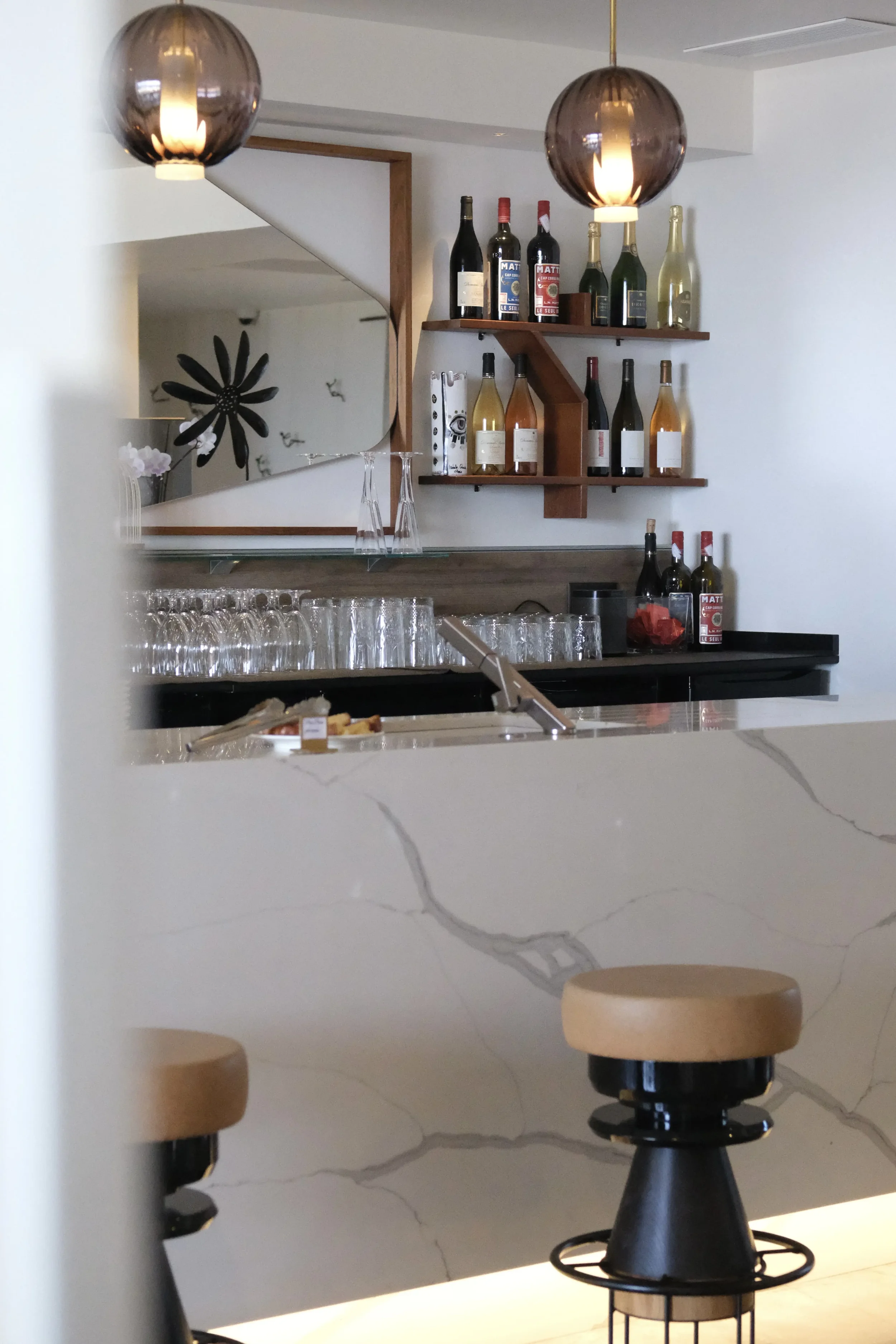 Un mur blanc avec une étagère en bois supportant des bouteilles de vin et de champagne, un grand miroir rectangulaire, deux chaises en bois avec assises en cuir, et un plan de travail en marbre beige avec des verres à vin empilés.