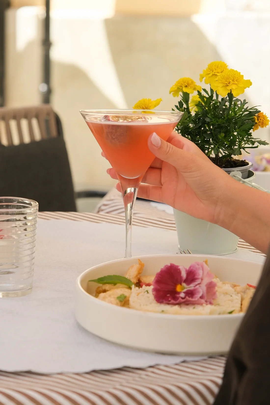 Une personne tient un verre à cocktail rose dans un repas en intérieur, avec un plat de nourriture, une plante avec des fleurs jaunes, et deux verres d'eau sur la table.