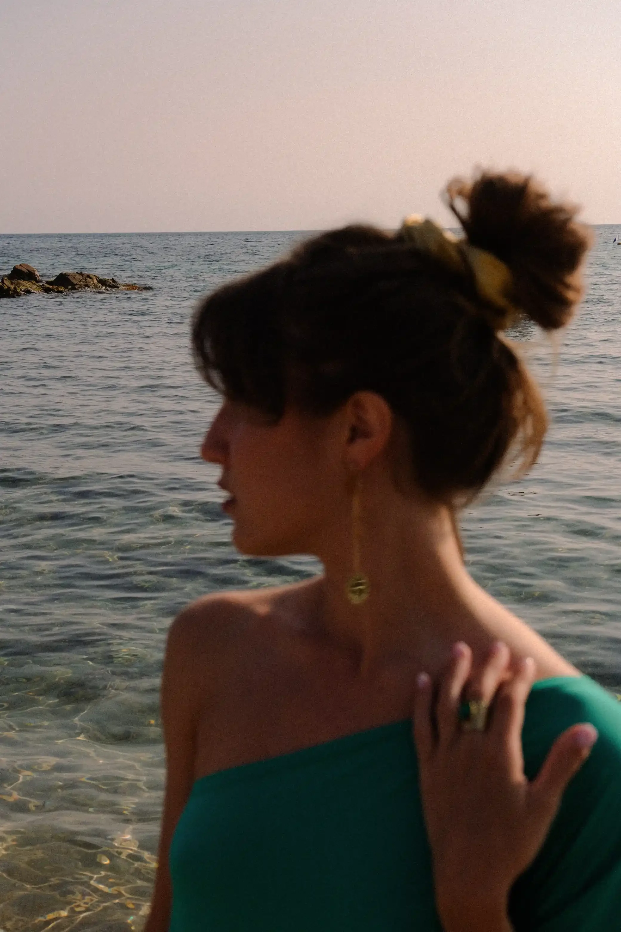 Une femme avec une coiffure en bun vue de profil, portant une boucle d'oreille, au bord de la mer avec des rochers et un bateau en arrière-plan.