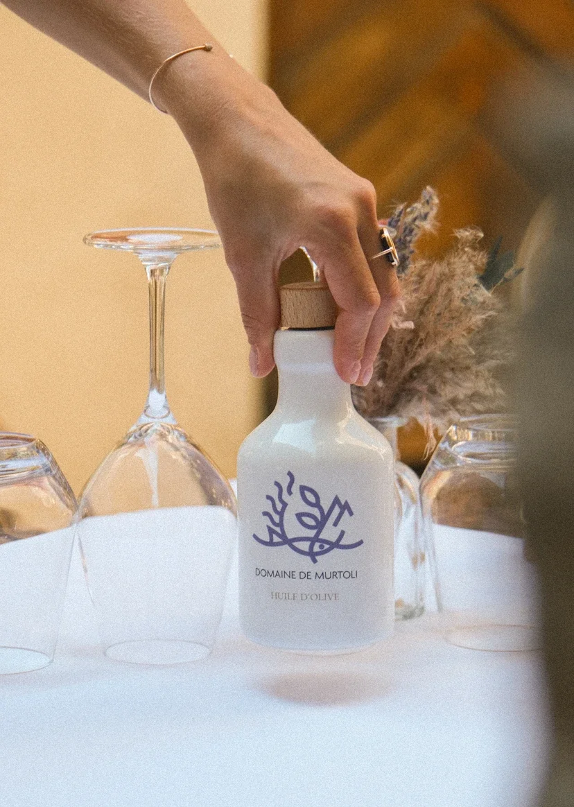 Main tenant une bouteille d'huile d'olive du domaine de Murtoli sur une table avec des verres à vin à l'envers.