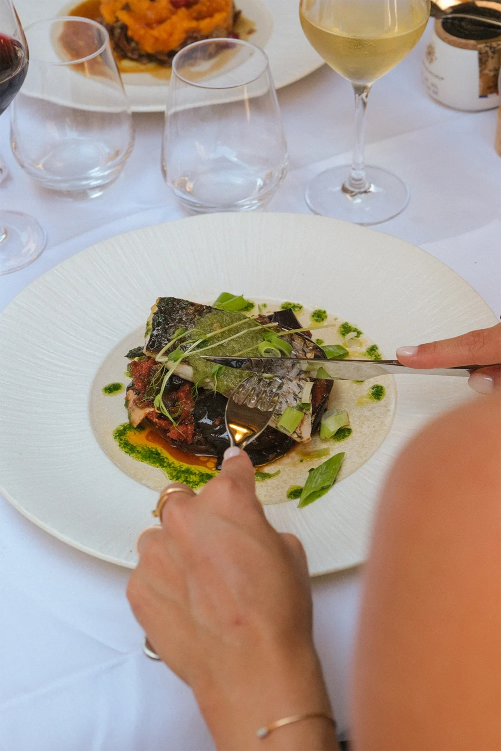 Une personne déguste un plat de poisson servi avec des légumes et une sauce verte, accompagnée de vin blanc, sur une table élégante.