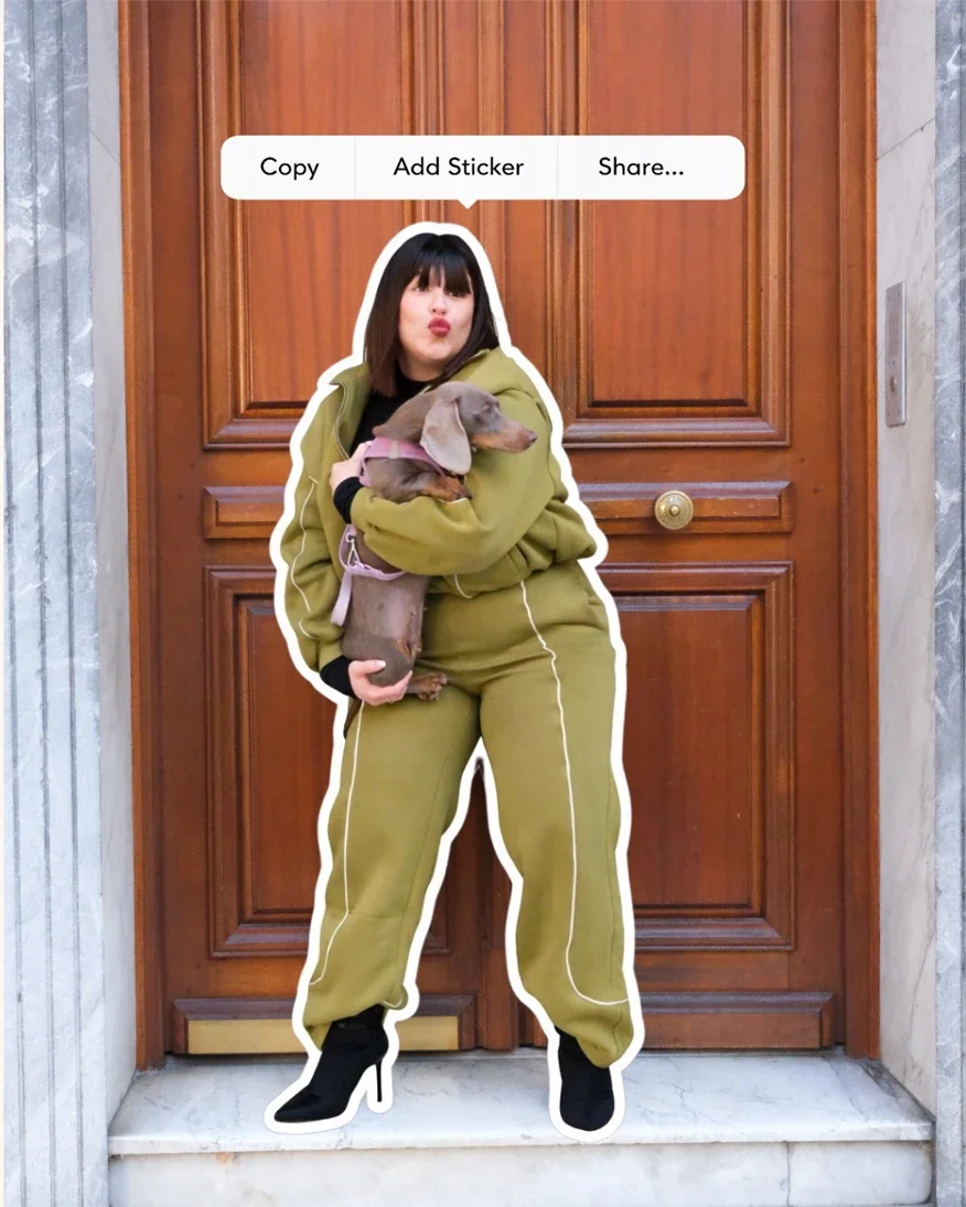 Une femme en sweat à capuche vert, portant des bottes à talons, tient un chien Dachshund. Elle fait une moue et se tient devant une porte en bois.