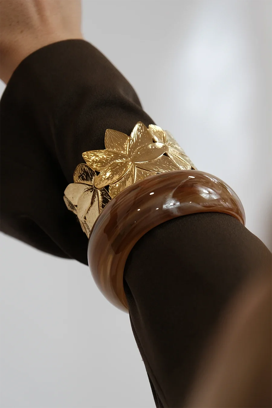 Poignet portant un bracelet en plastique marron et un bracelet orné de fleurs dorées, porté sur un vêtement noir.