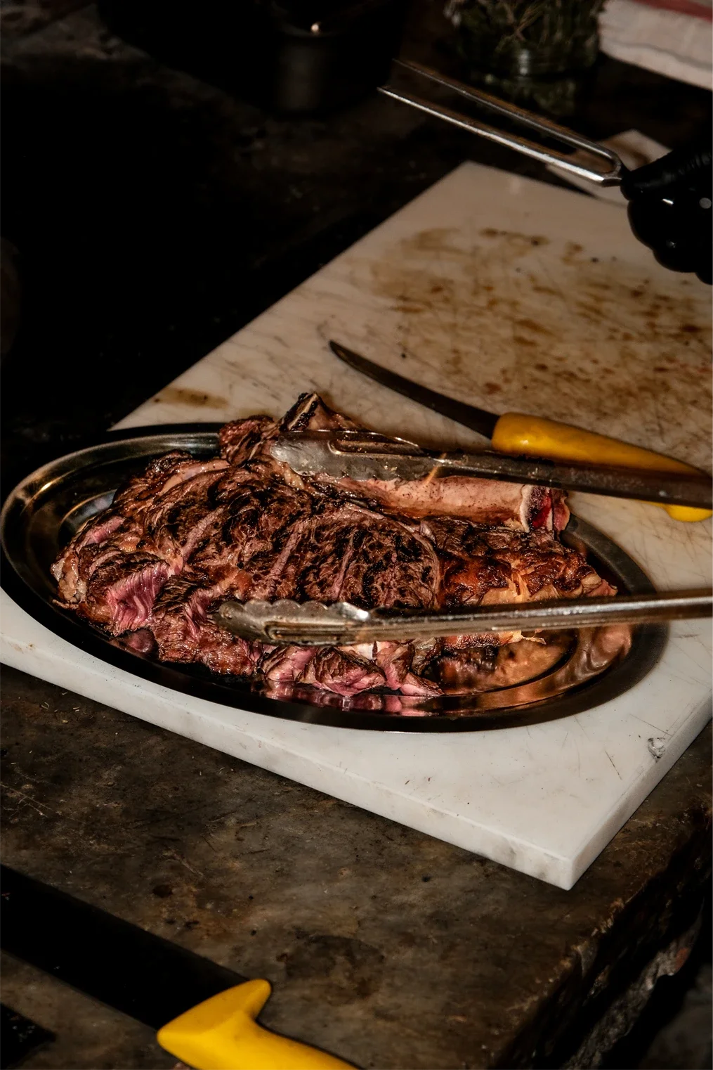 Une pièce de viande grillée sur une plaque en métal, avec des pinces à barbecue posées dessus, sur une surface en pierre ou en marbre. Un couteau avec une poignée jaune est à côté.
