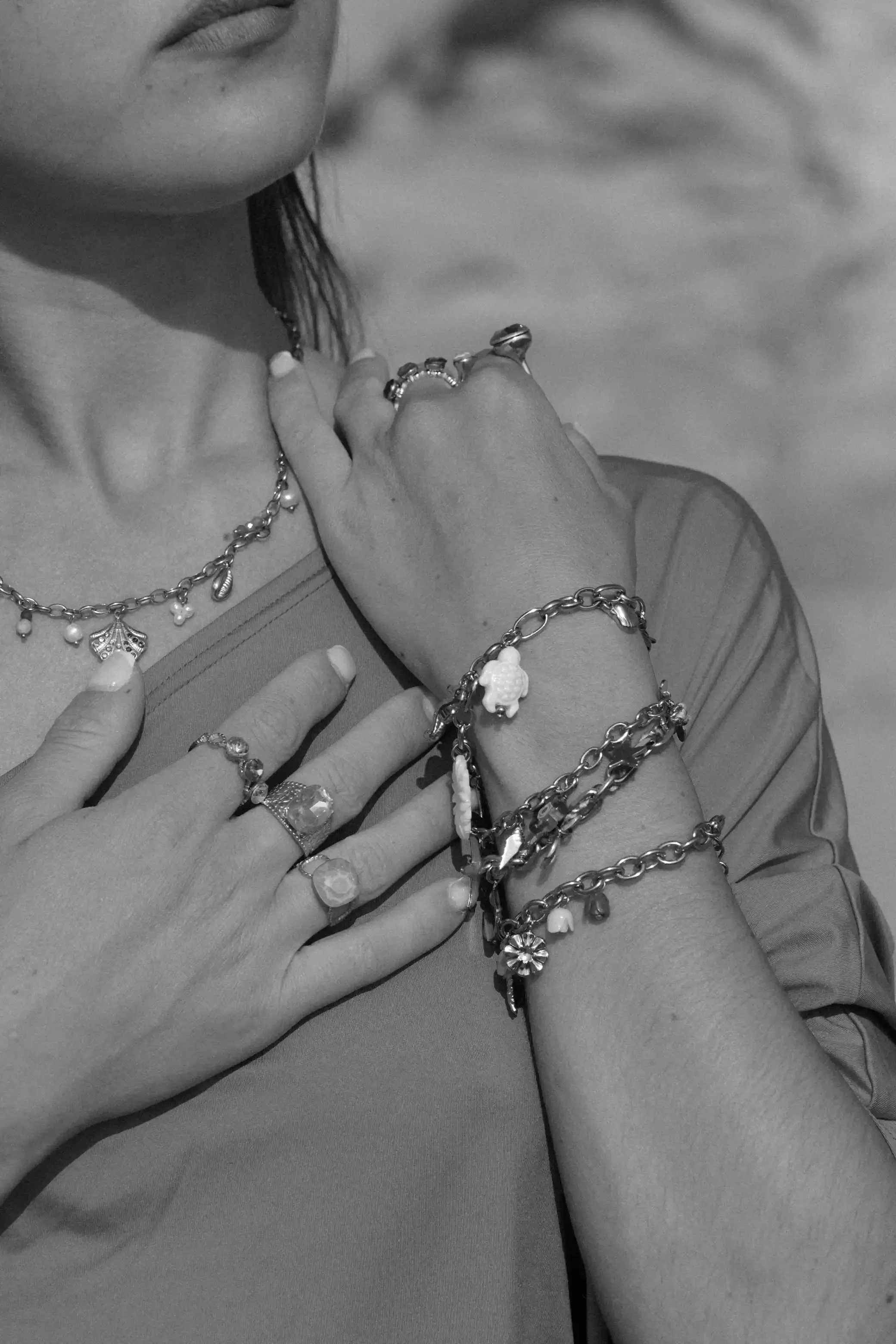 Première personne montrant ses mains, portant plusieurs bijoux en bracelets et bagues, avec un top à manches courtes, appareil photo en main, deuxième personne en arrière-plan également avec des bijoux. En noir et blanc.