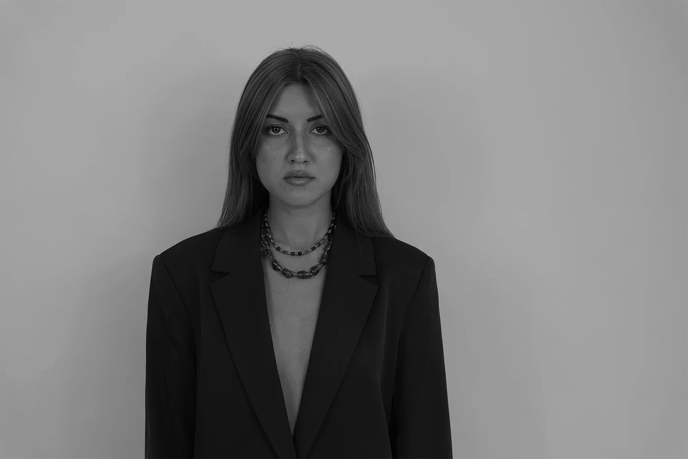 Portrait en noir et blanc d'une femme portant un blazer noir et plusieurs colliers, se tenant contre un mur clair.